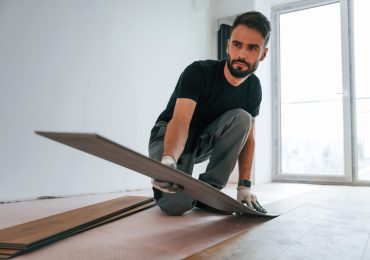 A focused man crouches on the floor, holding a plank of flooring, preparing to install it in a bright, empty room.