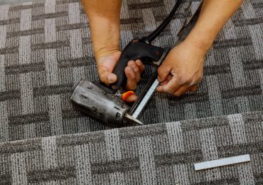 A person uses a power tool to cut carpet, hands focused on the task, with a textured gray carpet underneath.